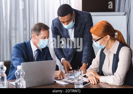 Des hommes d'affaires multiculturels dans des masques médicaux pointant du doigt vers un document sur le lieu de travail dans la salle de réunion Banque D'Images