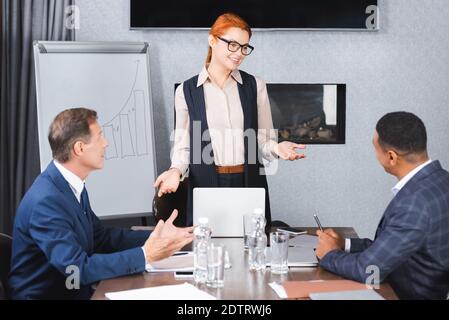 Sourire redhead exécutif gestant tout en parlant à des collègues multiculturels assis sur le lieu de travail pendant la réunion d'affaires Banque D'Images