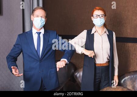 Hommes d'affaires dans des masques médicaux regardant l'appareil photo tout en saluant chacun autres avec coudes dans la salle de réunion Banque D'Images