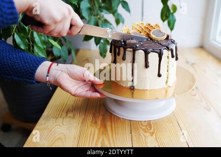 Les mains de la femme coupent le gâteau avec de la crème au caramel et de la ganache au chocolat sur une table en bois. Banque D'Images