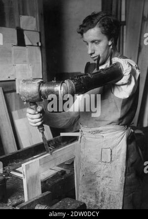 Ce jeune homme de 1921 ans utilise son bras prothétique brut du début du XXe siècle pour se faire vivre. À la place de son bras gauche, il a un club de cuir très brut, articulé qui est utile seulement pour frapper ou pousser des choses. La première Guerre mondiale a dévasté des millions de soldats et de familles. La légende de cette photo d'époque indique : « l'Allemagne essaie de reconstruire ses érables. La guerre a fait payer un lourd tribut à ce jeune homme, qui fait de son mieux dans la boutique du menuisier en dépit de ce handicap. Pour voir mes autres images médicales, recherche: Prestor vintage Medical Banque D'Images