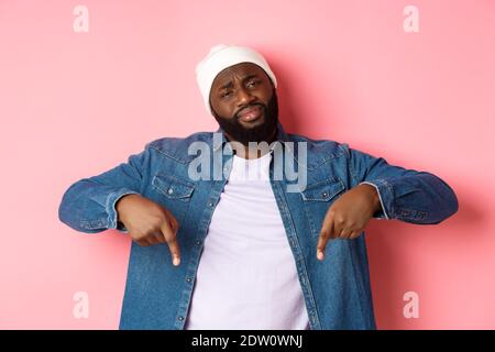 Sceptique et douteux Black Guy regardant la caméra non amusé, pointant les doigts vers le bas à quelque chose de mauvais, debout sur fond rose Banque D'Images