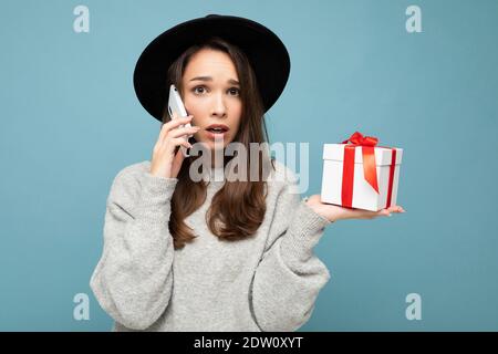 Plan de la jeune femme brunette assez triste choquée isolée le mur sur fond bleu porte un élégant chapeau noir et un chandail gris boîte cadeau en attente en conversation Banque D'Images