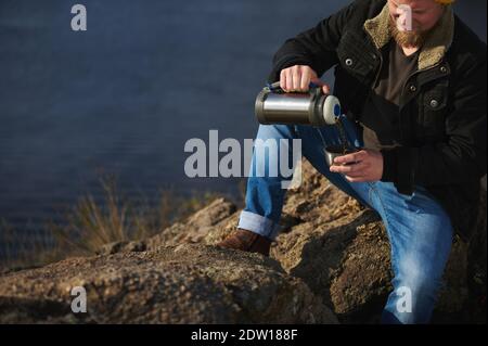 Portrait court d'un homme d'aventure assis sur des rochers tenant des thermos, verse une boisson chaude dans une tasse et fait une pause tout en randonnée dans la nature. Banque D'Images