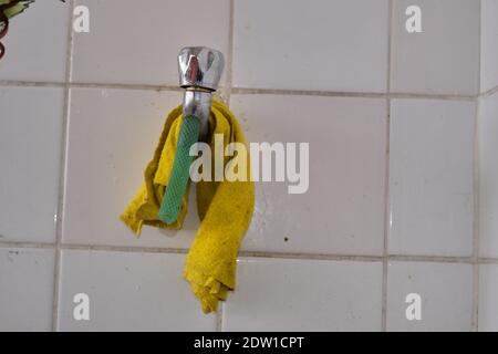Robinet en métal et bout de tuyau vert sur celui-ci. Serviette de nettoyage jaune accrochée à l'arrière-plan de la faïence blanche. Banque D'Images