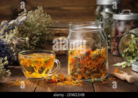 Tasse de thé marigold sain, pot en verre de fleurs calendula sèches. Pots d'herbes médicinales et livres anciens sur fond. Médecine alternative. Banque D'Images