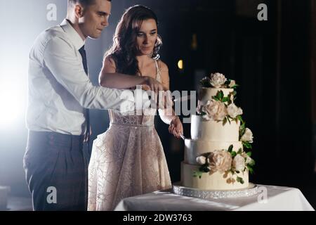 Des jeunes mariés concentrés coupent un gâteau de mariage à trois niveaux décoré de fleurs. Fête de mariage. Bonne mariée et marié Banque D'Images