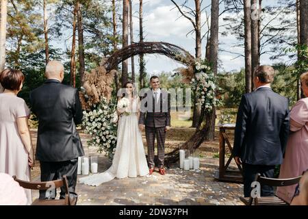 Mariée souriante et marié debout devant l'arche de mariage dans la forêt de pins. Heureux newlyweds à la cérémonie de mariage Banque D'Images
