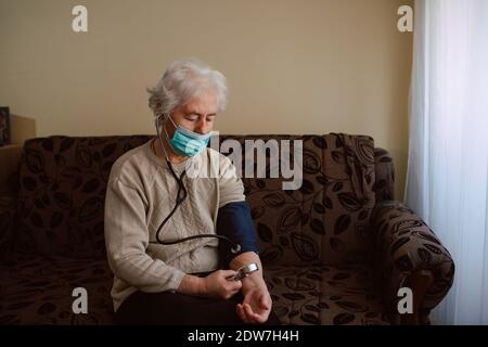 Femme caucasienne âgée avec un masque facial qui mesure la pression avec un manomètre tout en étant assis sur le canapé. Vérification de l'état de santé pendant le COVID-19 Banque D'Images