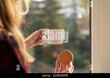 Une main femelle tient une tasse avec du café et des biscuits. Main de femme avec café et biscuits par la fenêtre comme arrière-plan avec place pour le texte Banque D'Images