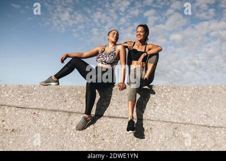 Deux amies assis dehors sur un mur parlant et souriant. femmes de fitness se reposant après l'entraînement sur un mur. Banque D'Images