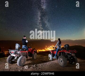 Vue arrière de couple heureux homme et femme touristes debout sur vtt quad motos sur le sommet de la montagne, appréciant la vue magnifique de ciel de nuit plein d'étoiles, voie lactée, village lumineux sur fond Banque D'Images