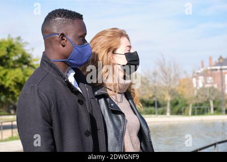 Homme noir africain et femme caucasienne portant des masques protecteurs dans un parc. Jeune couple multiracial pendant la pandémie du coronavirus. Banque D'Images