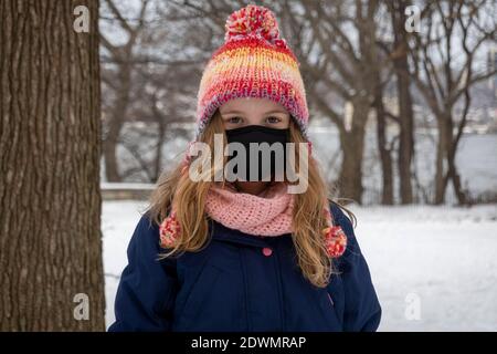 New York, États-Unis. Décembre 2020. Portrait d'une jeune fille portant un masque Covid et un chapeau de galet coloré debout dans la neige pendant l'hiver. Banque D'Images