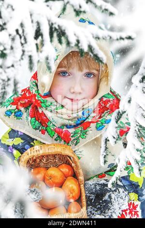 Une belle et heureuse fille aux yeux bleus dans un foulard brillant avec un panier de mandarines est assise sous un arbre de Noël enneigé. Carte de Noël Banque D'Images