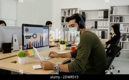 concept de distanciation sociale, homme d'affaires portant un masque, travaillant et regardant le papier graphique de la finance et le moniteur au bureau, s'asseoir loin des autres personnes Banque D'Images