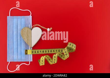 Concept de distanciation sociale - médaillon de coeur en bois de deux côtés séparés placés sur le masque médical à côté d'une mesure de ruban, tous isolés sur le dos rouge Banque D'Images