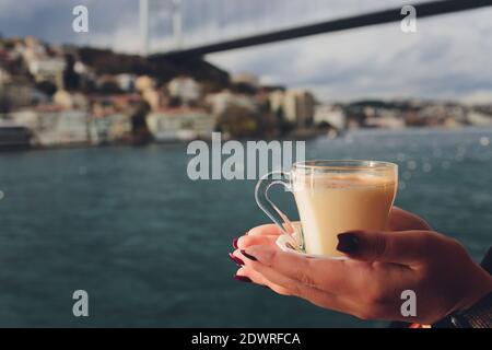 La main d'une femme tient une tasse blanche de chaud boisson lactée à la cannelle appelée sahlep de salep turc sur le Fond de l'eau ondulée et de la brumeuse Ma Banque D'Images