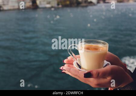 La main d'une femme tient une tasse blanche de chaud boisson lactée à la cannelle appelée sahlep de salep turc sur le Fond de l'eau ondulée et de la brumeuse Ma Banque D'Images