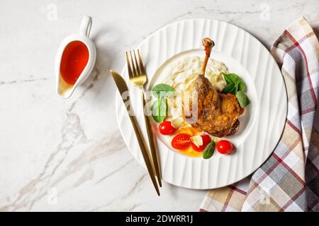 Cuisse de canard rôtie - confit de canard avec purée de panais et sauce à l'orange, tomates cerises, feuilles d'épinards fraîches servies sur une assiette blanche avec couverts dorés o Banque D'Images