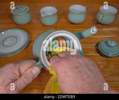 Gros plan de dessus de POV coup de tête des mains d'un homme en utilisant un torchon pour sécher une théière humide, sur un rainer de cuisine en pin, avec de la vaisselle connexe à proximité. Banque D'Images