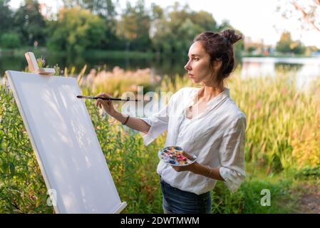 belle fille, femme artiste, en été sur le rivage du lac, étang de rivière, dessine l'image, dessin de début, premier coup de pinceau, toile blanche de pinceau Banque D'Images