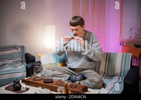 Un jeune homme goûte une tasse de thé chinois authentique fraîchement brassée dans un bol en verre. Plateau en bambou et plateau à thé sur la table Banque D'Images