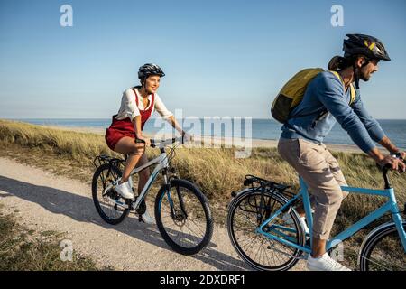 Jeune femme souriant en vélo avec un homme sur la piste de marche contre la mer Banque D'Images