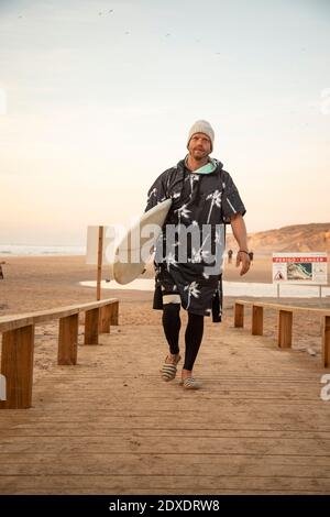 Homme adulte moyen transportant des planches de surf sur la promenade à la plage contre ciel Banque D'Images