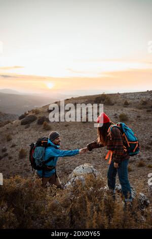Petite amie tenant la main d'un petit ami grimpant la montagne rocheuse contre le ciel au coucher du soleil Banque D'Images