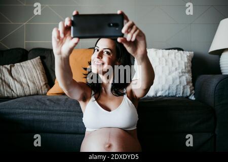 Femme enceinte souriante prenant le selfie dans le salon à la maison Banque D'Images