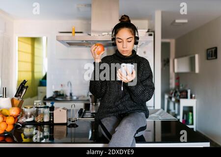 Adolescente portant un casque tenant une pomme tout en utilisant un téléphone portable à la maison Banque D'Images