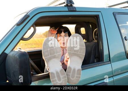 Surprise jeune femme assise avec les pieds sur la fenêtre de voiture au coucher du soleil Banque D'Images