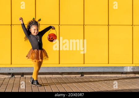 Fille souriante dansant avec pot de fleur d'Halloween et serre-tête contre paroi à motif coché jaune Banque D'Images