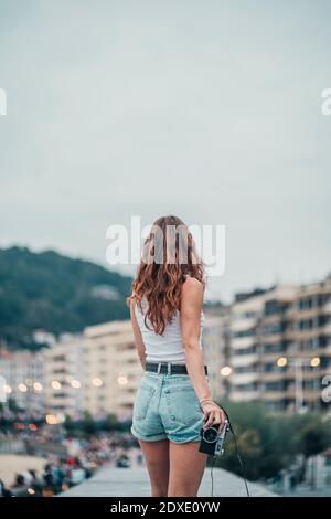 Jeune femme avec caméra regardant la vue sur la ville en se tenant debout contre le ciel Banque D'Images