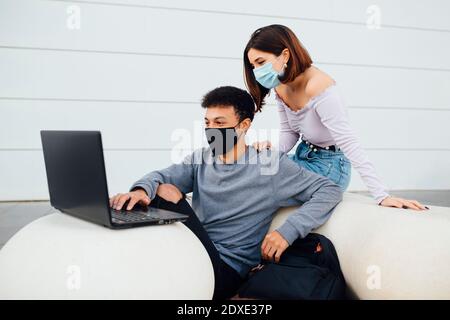Jeune couple portant un masque facial avec un ordinateur portable assis près boule en béton blanc contre le mur Banque D'Images