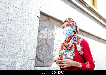 Femme en hijab portant un masque de protection tenant un smartphone Tout en marchant par le mur pendant COVID-19 Banque D'Images