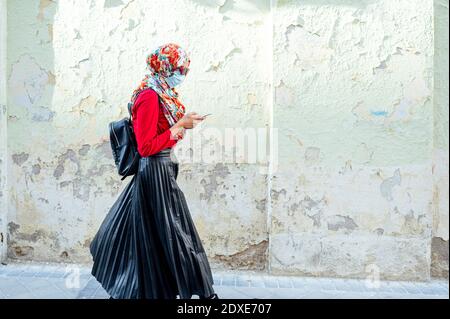 Femme musulmane portant un masque de protection sur un téléphone portable pendant marcher par le mur pendant le coronavirus Banque D'Images