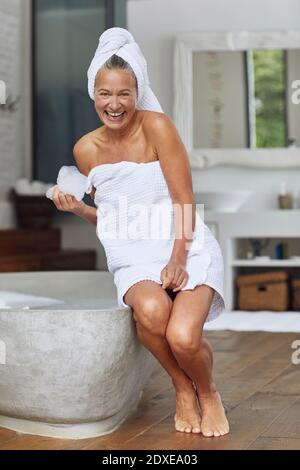 Bonne femme mûre jouant avec des bulles tout en étant assise sur le bord de baignoire dans la salle de bains Banque D'Images