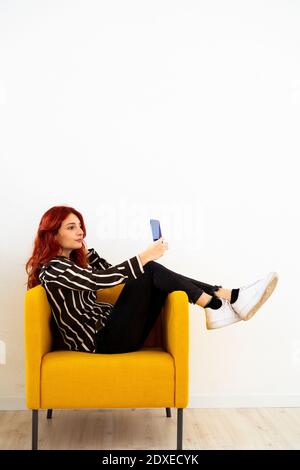 Une jeune femme prend le selfie par téléphone portable tout en étant assise fauteuil au bureau Banque D'Images