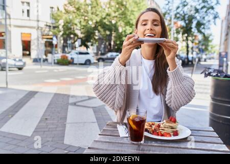 Femme prenant la photo de la nourriture et des boissons sur la table pendant assis sur le trottoir au café Banque D'Images