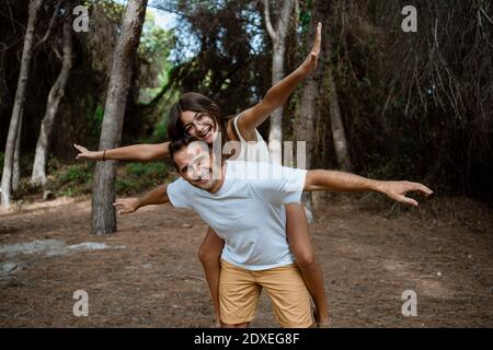 Un père souriant donne une promenade en porcgyback à sa fille avec les bras étirés en forêt pendant les vacances Banque D'Images