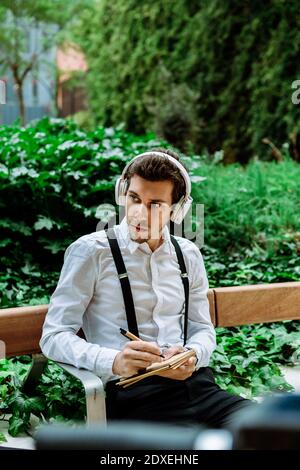 Jeune homme d'affaires avec casque assis sur un banc à l'extérieur et à l'écriture dans le bloc-notes Banque D'Images