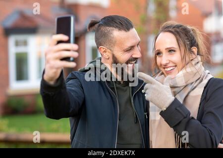 Jeune femme en train de gesturer tout en prenant le selfie avec un ami gai dehors nouvelle maison Banque D'Images