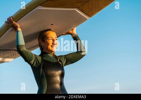 Femme adulte portant un paddleboard sur la tête tout en se tenant contre le bleu ciel Banque D'Images