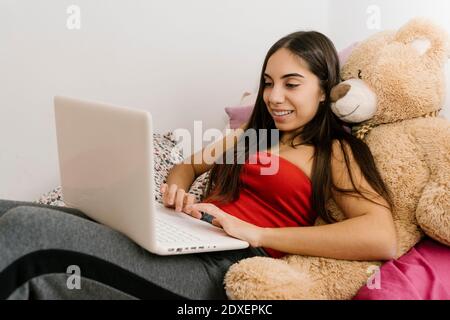 Adolescente souriante utilisant un ordinateur portable tout en se penchant sur l'ours en peluche jouet à la maison Banque D'Images