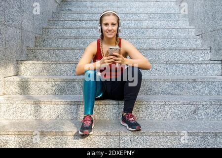 Sportswoman avec une jambe prothétique souriant tout en utilisant le téléphone mobile assis sur les marches Banque D'Images