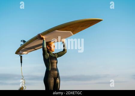 Femme mature regardant loin en portant le paddleboard debout contre le ciel bleu Banque D'Images