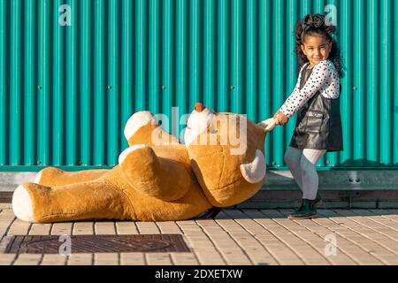 Une fille traînant un ours en peluche sur le sentier tout en se tenant contre le vert mur Banque D'Images