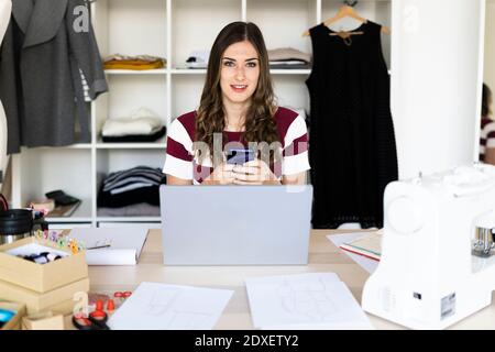 Jeune femme d'affaires tenant un smartphone assis avec un ordinateur portable à studio Banque D'Images